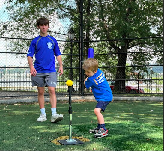 Little boy batting at tee while counselor watches